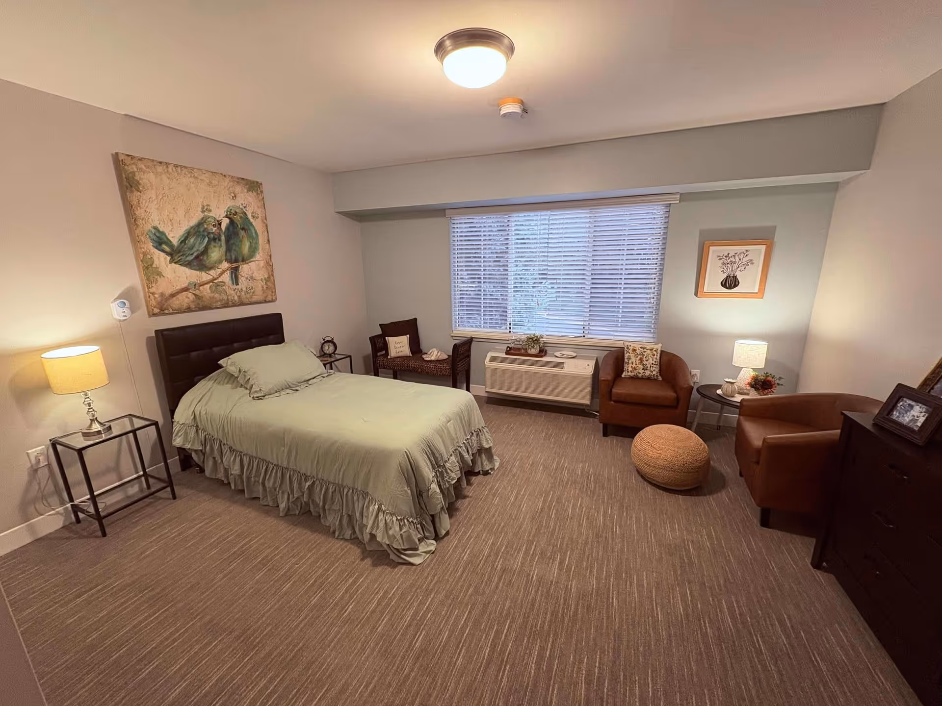 A cozy bedroom in a senior living facility featuring a single bed with a green bedspread, a nightstand with a lamp, two brown armchairs, a small round table with a lamp and decorative items, a cushioned bench, a large window with blinds, and artwork on the walls.