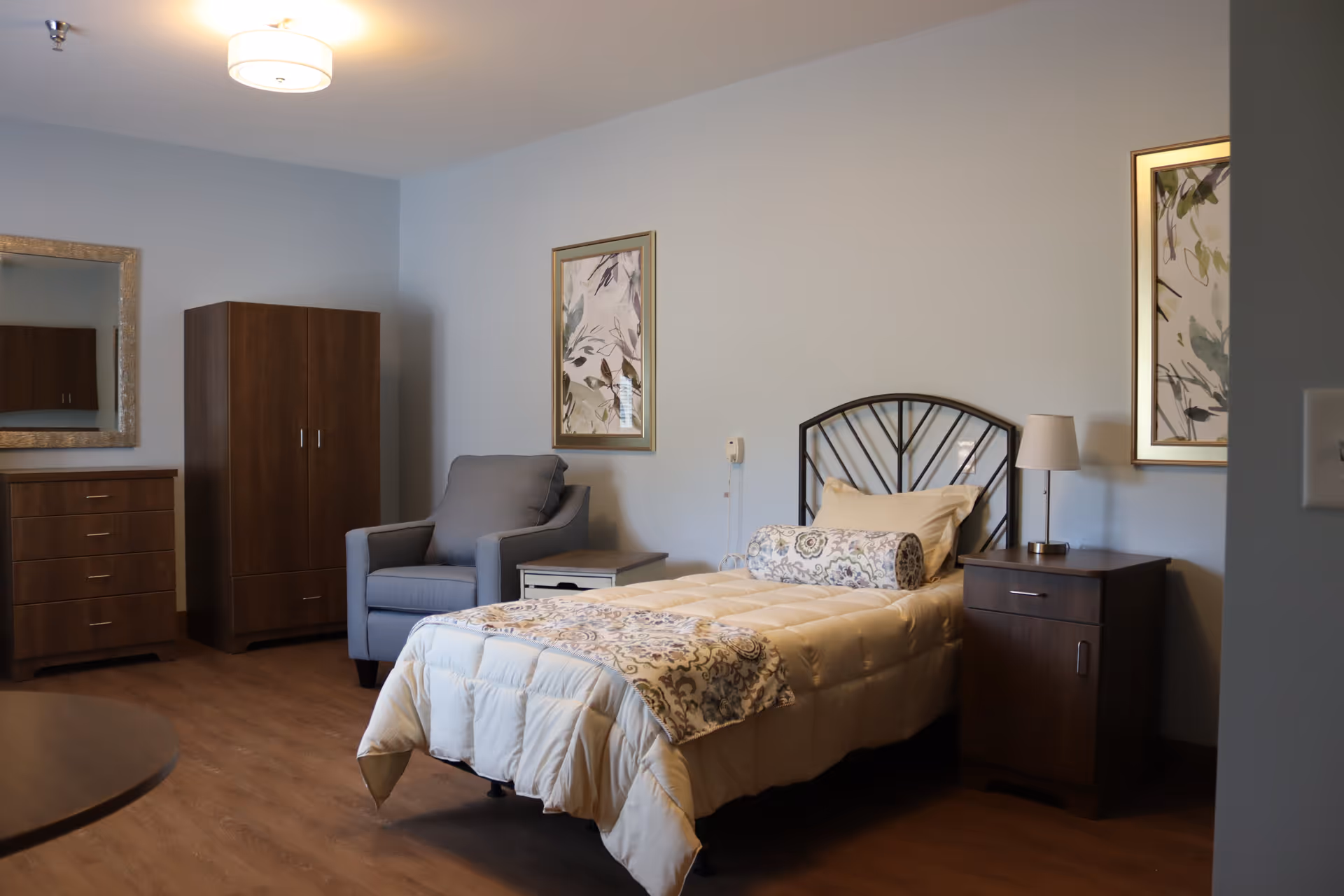 A furnished assisted-living bedroom with a single bed, nightstand and lamp, upholstered armchair, wardrobe and dresser against a pale blue wall.