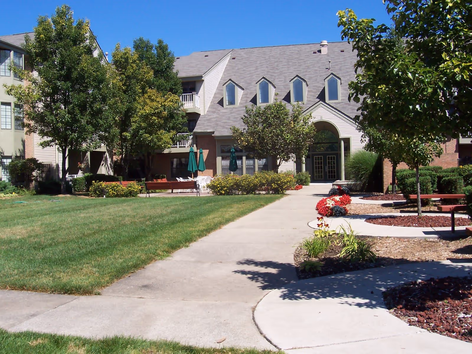 Exterior view of Rosehaven Manor showing a multi-story building with a gray roof and several windows. The foreground features a well-maintained lawn, a concrete walkway, trees, flower beds with red and white flowers, benches, and outdoor seating with green umbrellas under a clear blue sky.