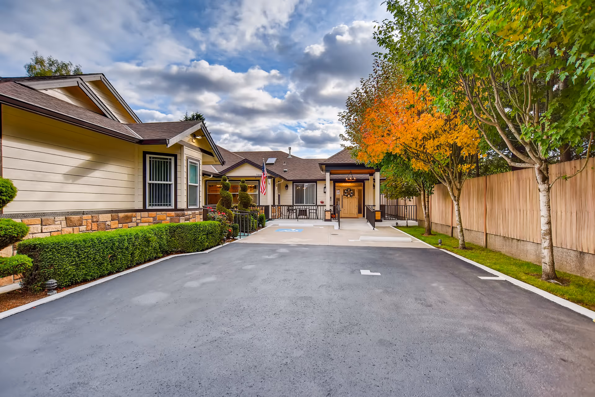 View of the front entrance of a single-story senior living facility with a paved driveway, manicured bushes, and trees with autumn foliage under a partly cloudy sky.
