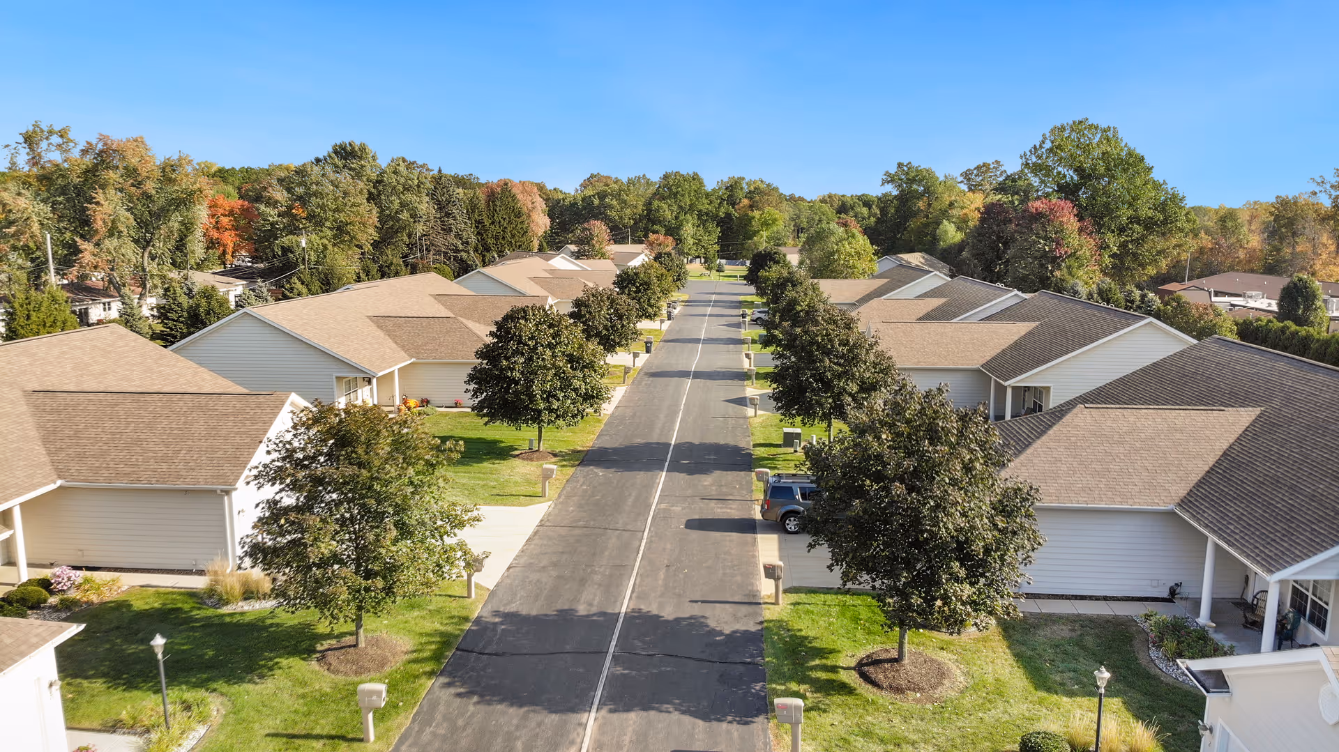 Aerial view of a residential street in a senior living community with single-story houses on both sides, green lawns, trees, and a clear blue sky.