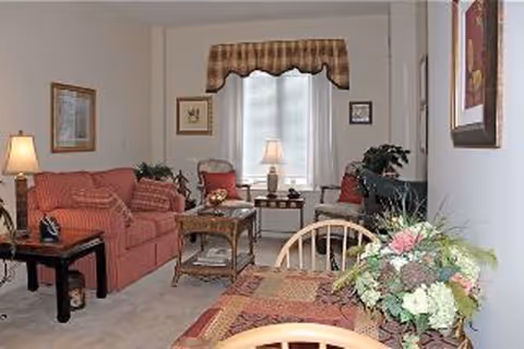 A cozy living room area with a red sofa, a wooden coffee table, a side table with a lamp, and a window with a valance. There is a dining table with a floral centerpiece in the foreground and framed artwork on the walls.