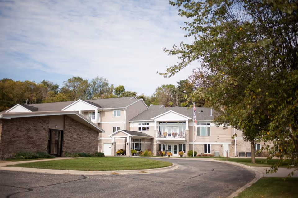 Exterior view of a multi-story assisted living facility with beige siding and brick accents, surrounded by trees and greenery under a partly cloudy sky. A curved driveway leads to the main entrance with a covered porch and an American flag on a flagpole nearby.