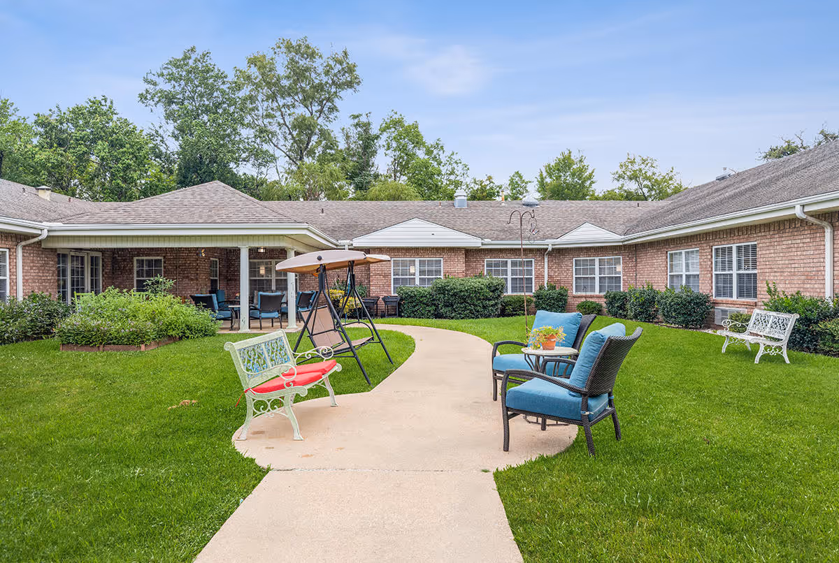 Outdoor courtyard area of Broadmoor Court senior living facility featuring a paved walkway, green lawn, patio swing, benches with cushions, and chairs with a small table. The building surrounds the courtyard with brick walls and multiple windows. Trees and bushes are visible in the background under a clear sky.