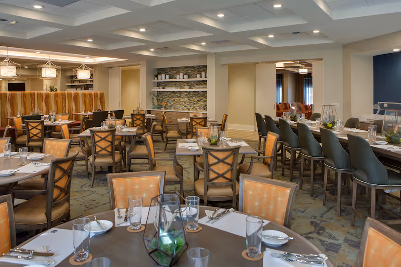 A spacious dining room with multiple tables and chairs arranged neatly. Tables are set with plates, glasses, and cutlery. The room features warm lighting with pendant lights hanging from the ceiling, a patterned carpet, and a decorative wall with shelves holding plates and plants.