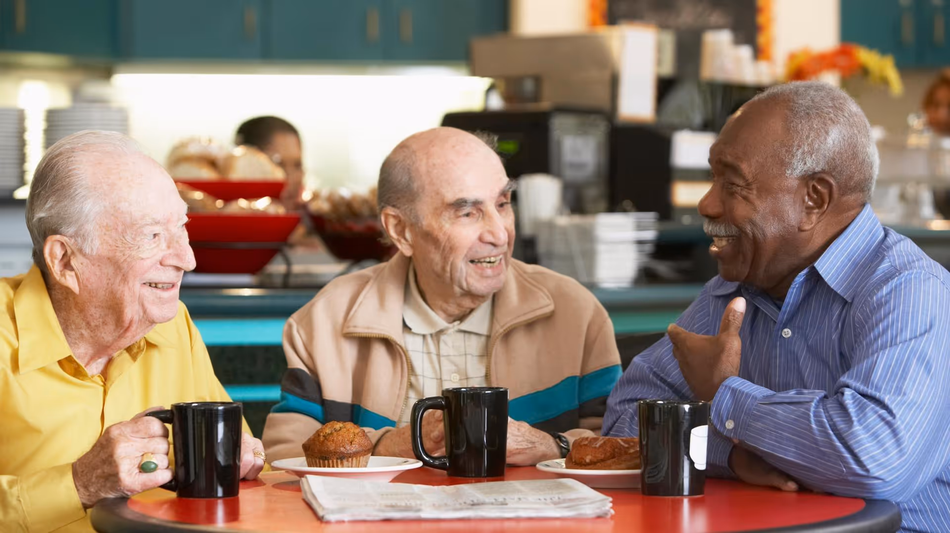 Three elderly men sitting around a table in a dining area, smiling and engaging in conversation. Each has a black coffee mug and a plate with a muffin or pastry in front of them. The background shows a kitchen or cafeteria setting with dishes and food items.