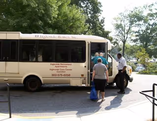 A small beige shuttle bus labeled Ingleside Homes Inc is parked on a driveway. Several elderly people are boarding the bus with assistance from a staff member. Trees and parked cars are visible in the background.
