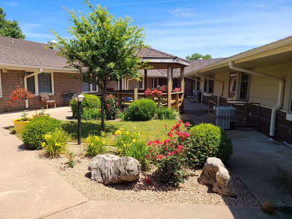 Outdoor courtyard area at a senior living community featuring a small garden with bushes, flowers, rocks, and a tree. There is a wooden gazebo in the center surrounded by a paved walkway and brick buildings with windows and chairs along the sides under a clear blue sky.