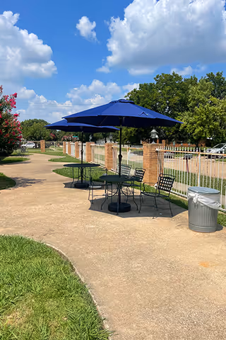 Outdoor paved walkway with black metal tables and chairs shaded by large blue umbrellas, bordered by a white fence and brick pillars, with green grass and trees under a partly cloudy blue sky.