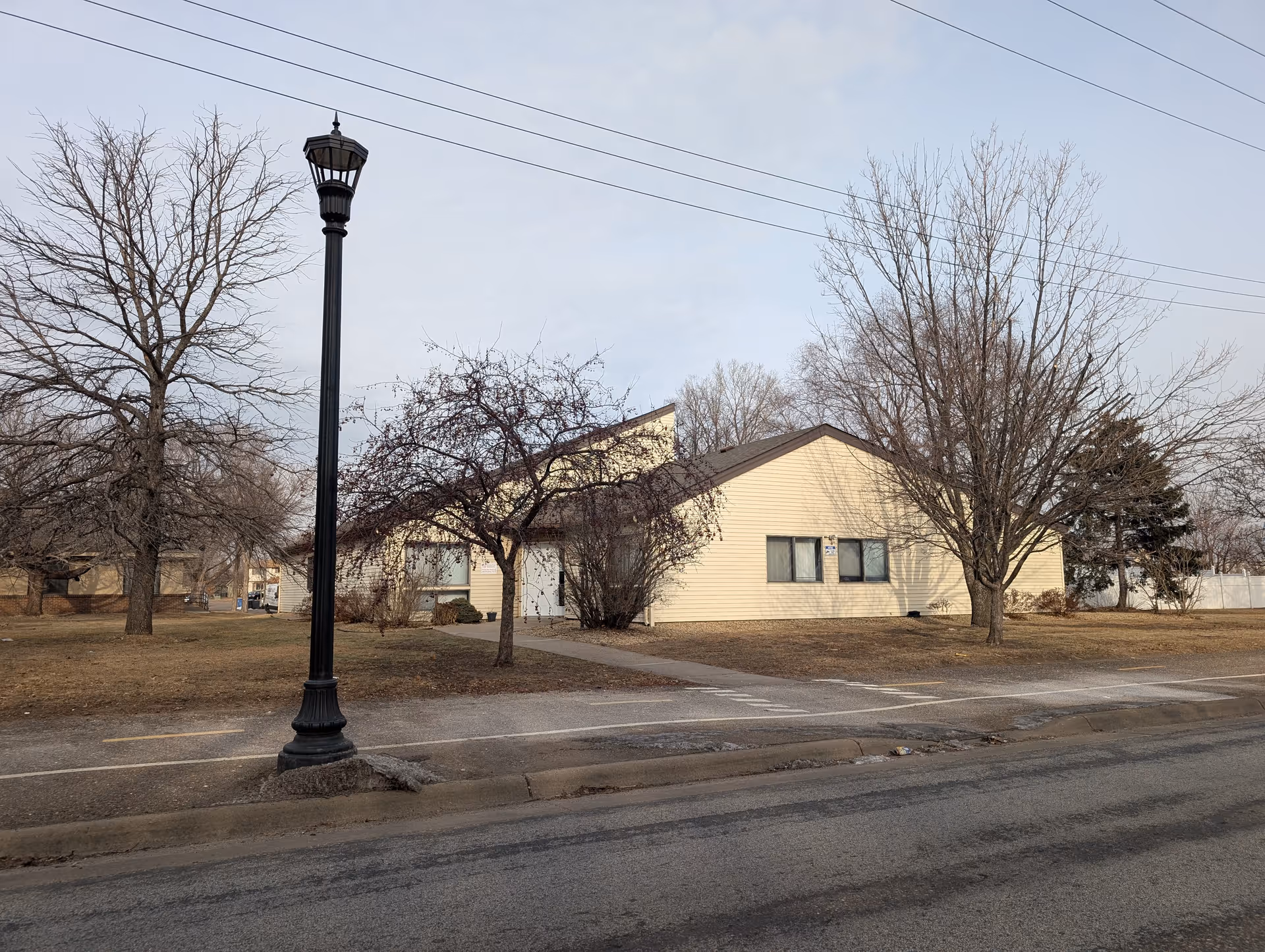 Exterior view of a single-story beige building with a slanted roof, surrounded by leafless trees and a sidewalk. A black street lamp is visible in the foreground, and the sky is overcast.
