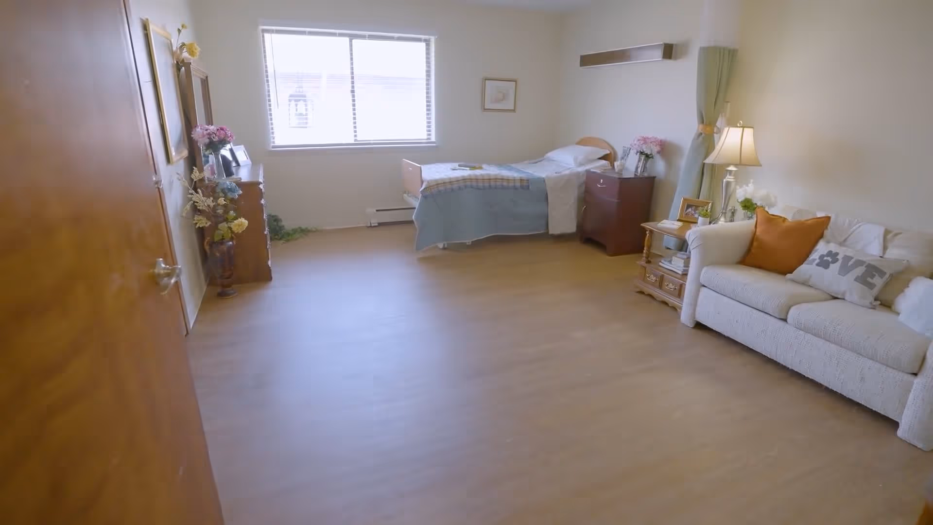A bright and spacious assisted living bedroom with a single bed covered with a blue blanket and white sheets, a wooden nightstand with flowers and framed photos, a beige couch with decorative pillows including one with the word 'LOVE', a wooden side table with a lamp and more framed photos, and a large window letting in natural light.