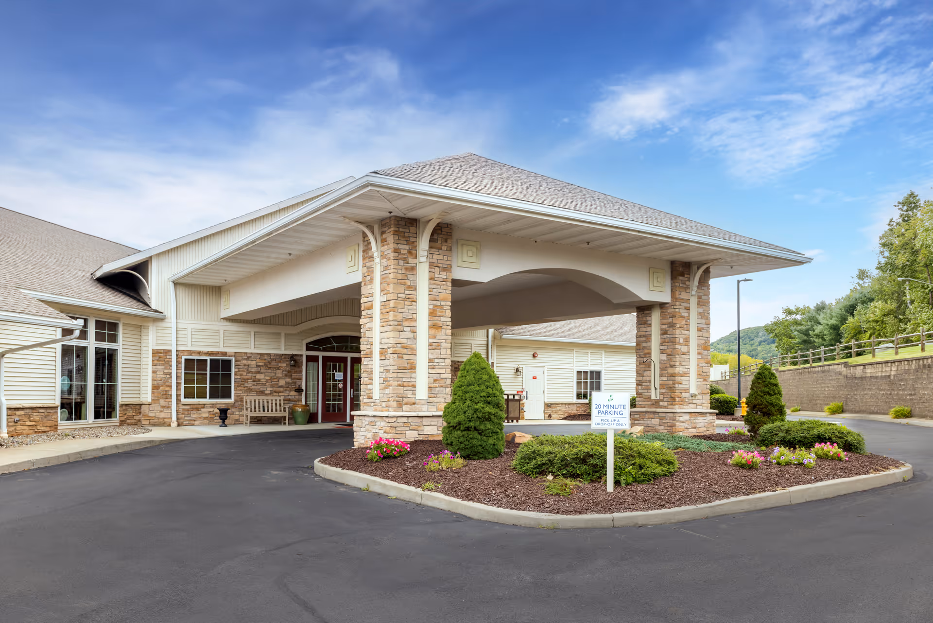 Entrance of a senior living facility with a covered driveway supported by stone pillars. There is a landscaped area with bushes and flowers in front, and a sign indicating 20 minute parking for pick-up and drop-off only. The building has beige siding and large windows, with a clear blue sky in the background.