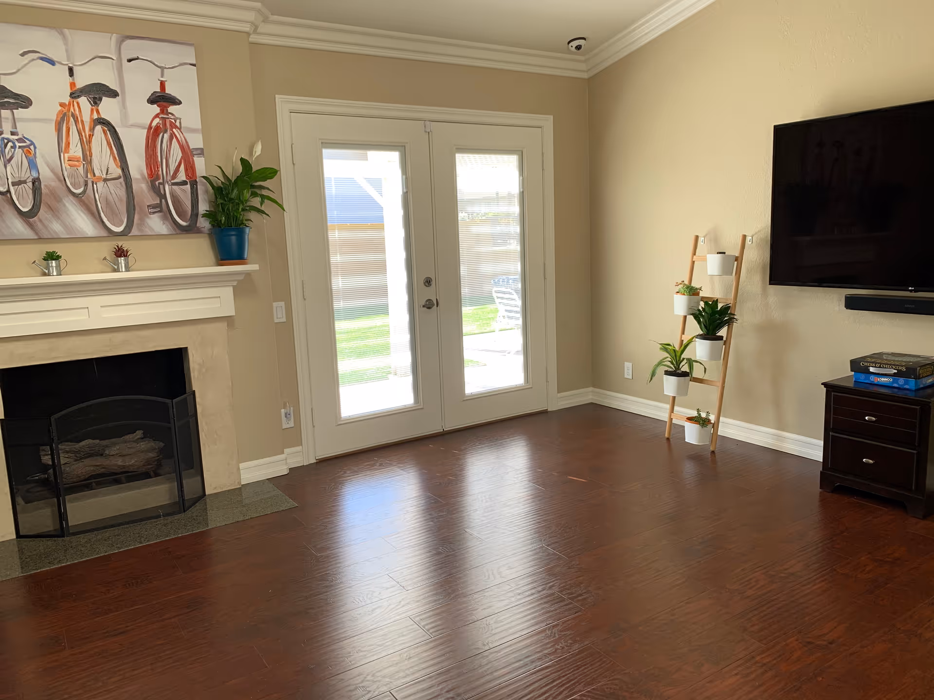 A bright living room with wooden flooring, a fireplace with a black screen, a painting of three bicycles above the fireplace, a set of double glass doors leading outside, a wooden ladder shelf with potted plants, a wall-mounted flat-screen TV, and a small dark wooden cabinet with board games on top.