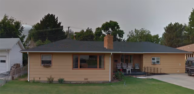 Single-story beige house with a dark roof, a chimney, and a front porch with chairs. There is a green lawn in front and trees in the background under a cloudy sky.