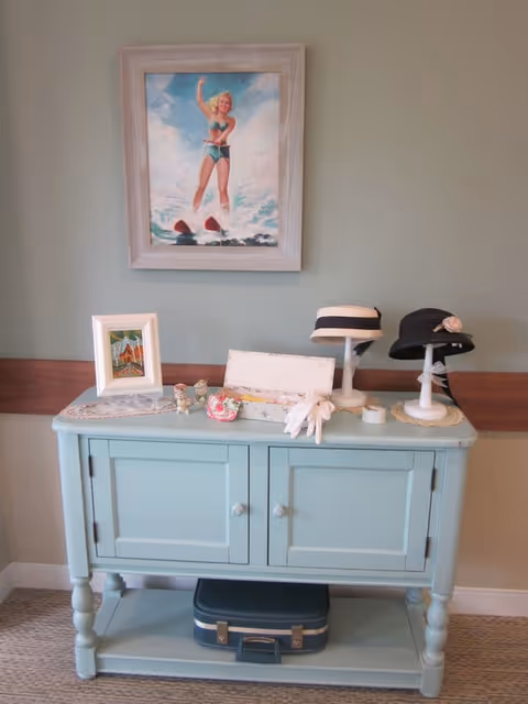A light blue wooden cabinet with two doors and a lower shelf holding a vintage suitcase. On top of the cabinet are decorative items including two hats on stands, a pair of white gloves, small figurines, a framed picture, and a painting of a woman water skiing hanging on the wall above.