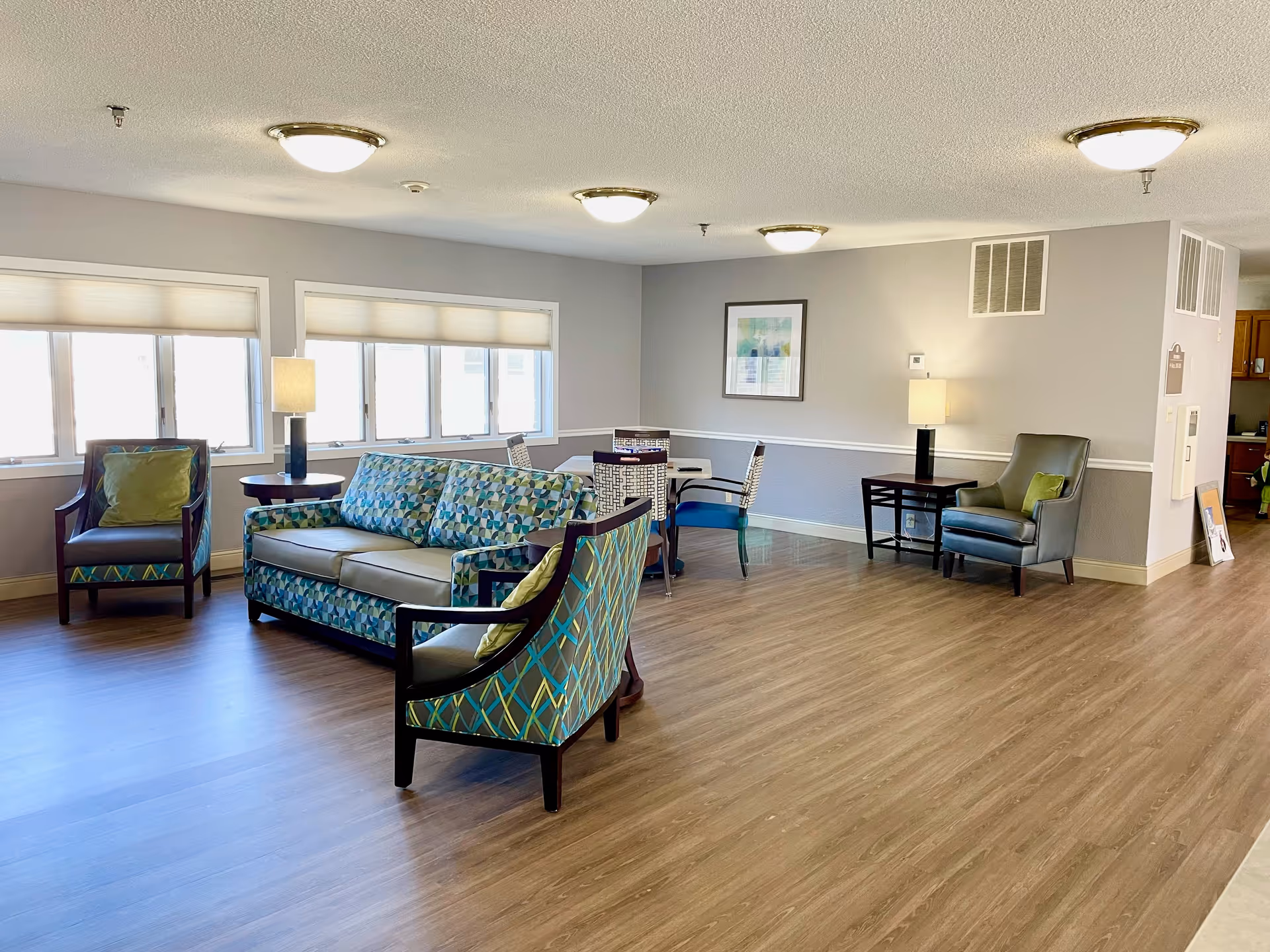 A bright and spacious senior living community common area with wooden flooring, patterned upholstered sofa and chairs, side tables with lamps, a small table with chairs, and large windows letting in natural light.