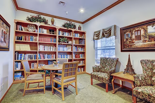 A cozy reading room with a large wooden bookshelf filled with books and decorative plants on top. There is a round wooden table with four chairs in the center of the room. Two upholstered armchairs with floral patterns are placed near a window with a valance. A small wooden side table between the armchairs holds a decorative Eiffel Tower sculpture. The walls are adorned with framed paintings.