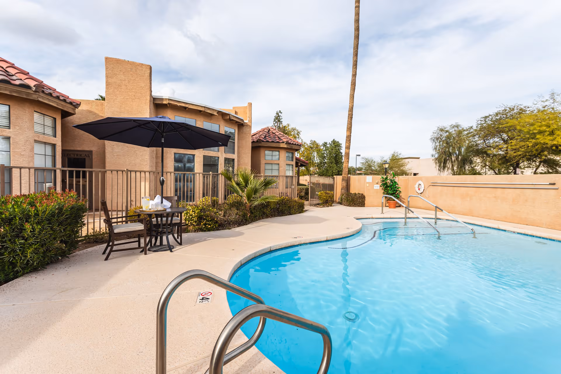 Outdoor swimming pool with metal handrails, a patio table and umbrella, and a stucco building in the background.