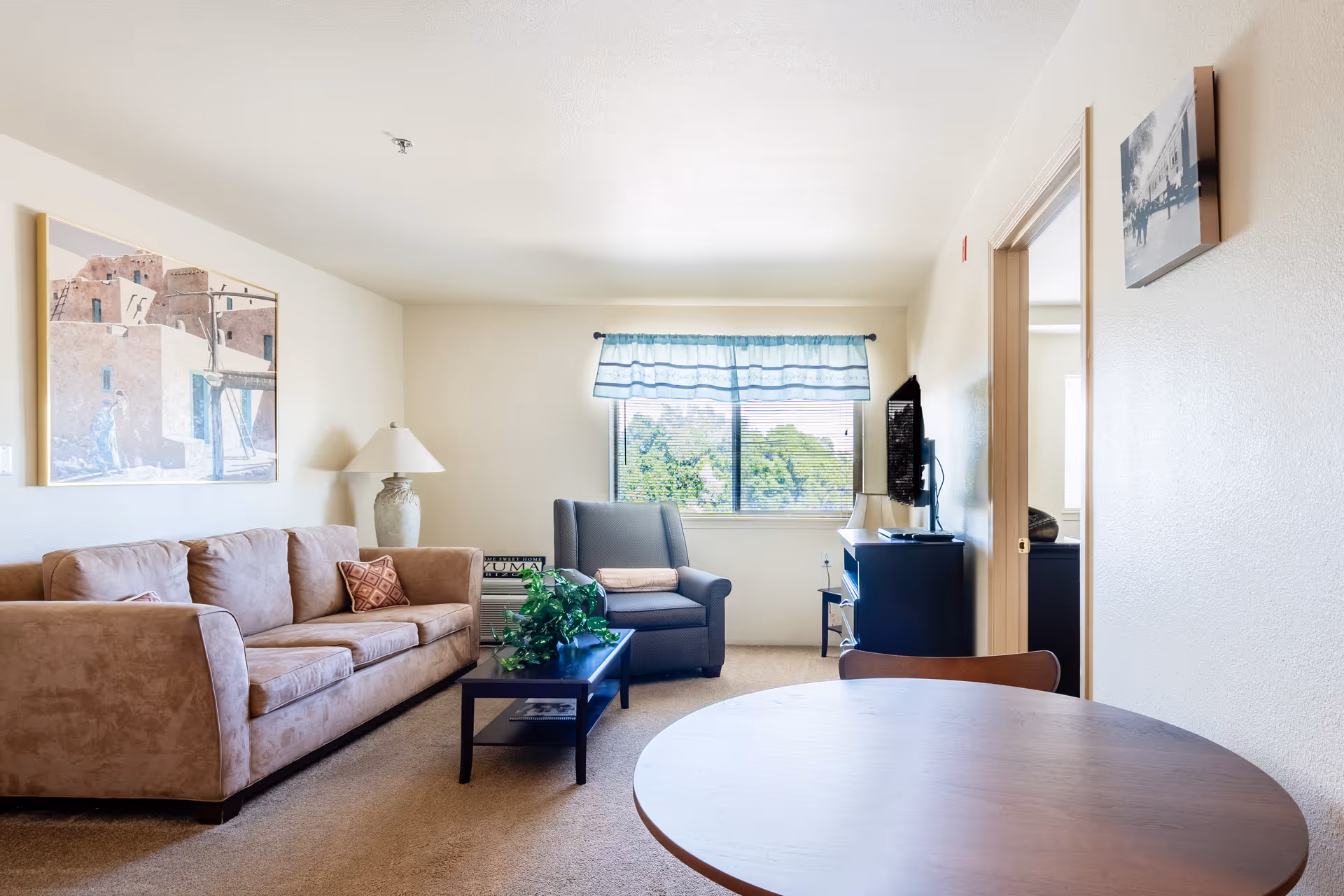A bright living room in a retirement community featuring a beige sofa with a decorative pillow, a large lamp on a side table, a dark armchair with a folded blanket on it, a coffee table with a green plant, a window with blue valance curtains, a wall-mounted TV, and a round wooden table in the foreground. There is a doorway leading to another room and artwork on the walls.