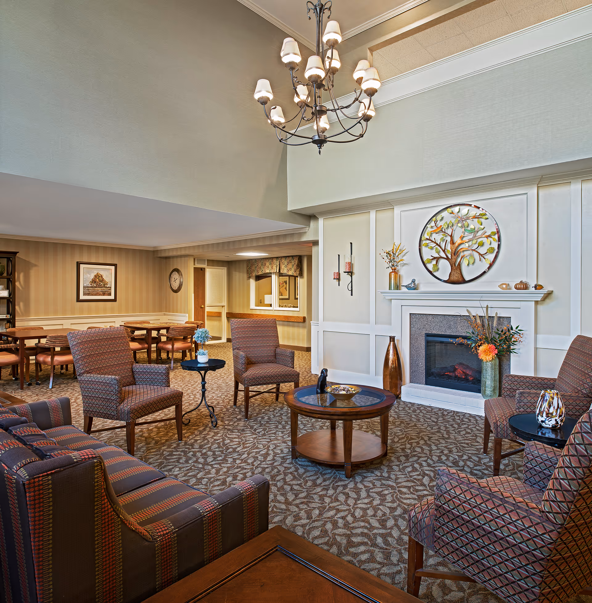 Comfortable senior living common room with upholstered chairs and sofas arranged around coffee tables and a fireplace under a chandelier.