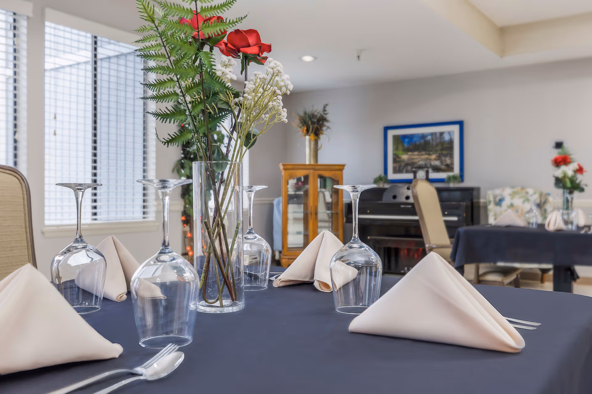 A dining table set with upside-down wine glasses, folded beige napkins, and a clear vase holding artificial flowers including a red rose and white baby's breath. In the background, there are more tables with similar settings, a piano, a wooden cabinet with glass doors, and a framed picture on the wall.