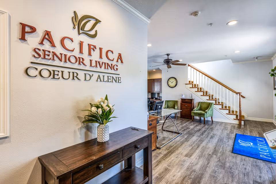 Interior view of a senior living facility reception area with a wooden table holding a vase of white flowers, a wall sign reading 'Pacifica Senior Living Coeur d'Alene', two green chairs, a clock on the wall, a staircase with wooden handrails, and a blue floor mat with the Pacifica logo.