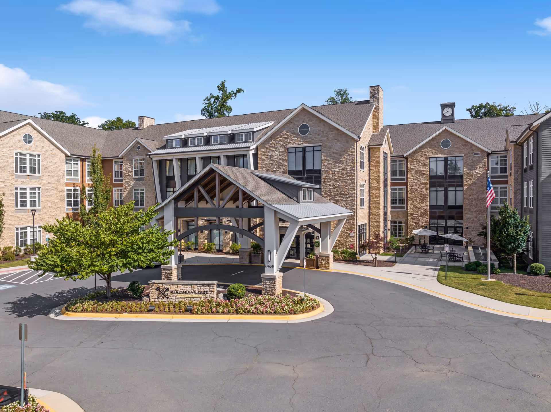 Exterior view of Heritage Village Assisted Living and Memory Care building with a covered entrance, stone facade, multiple windows, a clock tower, an American flag, and landscaped surroundings under a clear blue sky.
