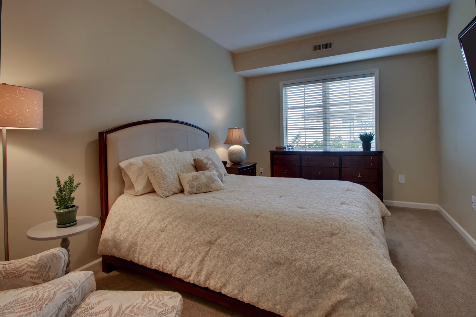 A cozy bedroom with a large bed covered in a beige patterned comforter and multiple pillows. There is a wooden headboard, a nightstand with a lamp, a small round table with a potted plant, and an upholstered chair. A large window with white blinds lets in natural light, and a dark wooden dresser is placed beneath the window.
