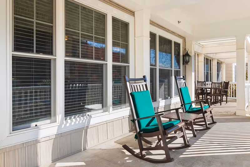 A covered outdoor patio area with two wooden rocking chairs featuring green cushions in the foreground and a wooden table with chairs in the background, adjacent to large windows with white frames.