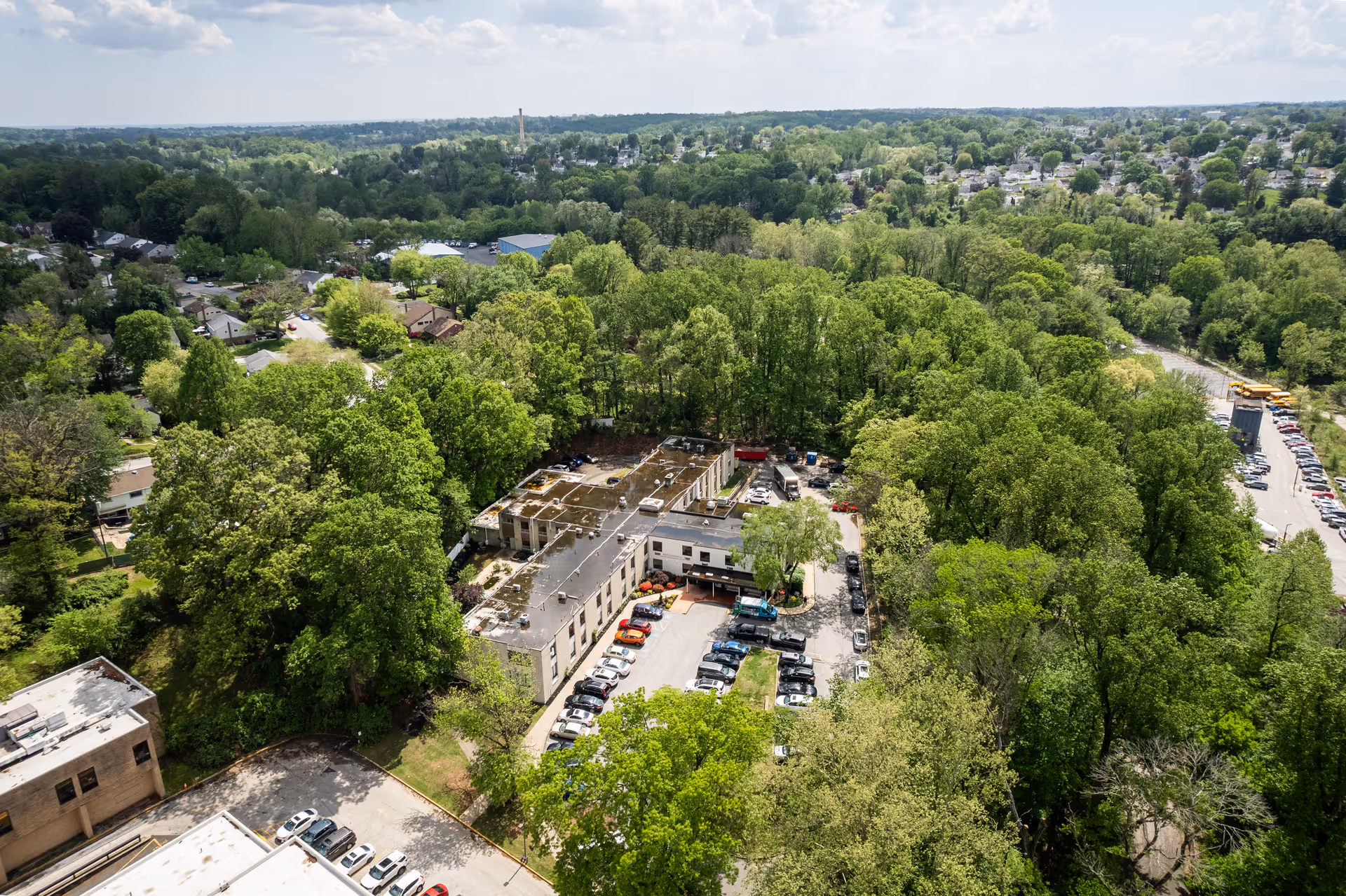 Aerial view of Westgate Hills Rehabilitation & Nursing Center surrounded by dense green trees and residential neighborhoods. The building is a two-story structure with a parking lot filled with cars. The area is lush with greenery and the horizon shows more trees and houses under a clear sky.