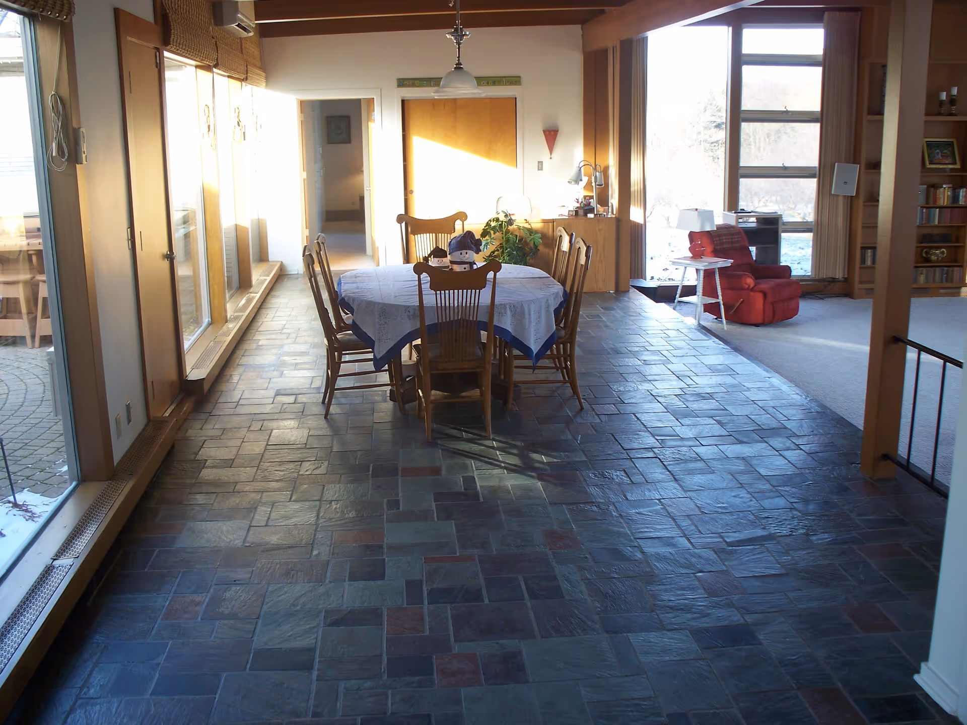 Interior view of a senior living facility dining area with a round table covered with a white and blue tablecloth surrounded by six wooden chairs. The floor is tiled with multi-colored stone tiles. To the right, there is a red recliner chair next to a small white side table with a lamp. Large windows let in natural light, and a bookshelf is visible in the background.
