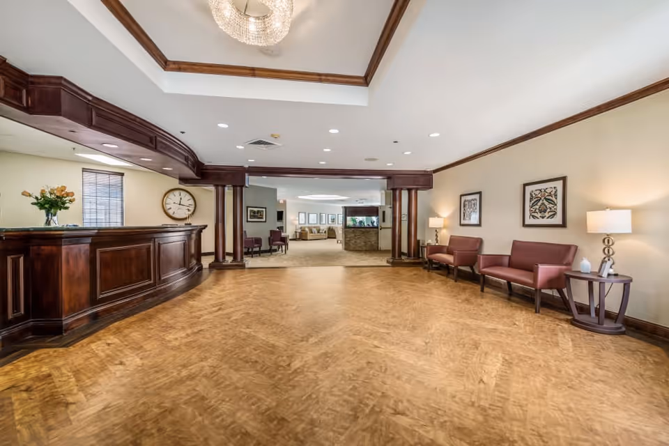 Spacious reception area with a wooden front desk on the left, a large clock on the wall, and a seating area with maroon chairs and side tables with lamps on the right. The room has wood trim accents and a chandelier hanging from the ceiling. In the background, there is an open area with additional seating and framed artwork on the walls.