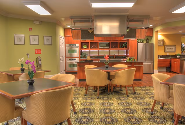 A spacious kitchen and dining area with green walls and patterned carpet. The kitchen features wooden cabinets, stainless steel appliances including a double oven and refrigerator, and a large range hood. Several tables with beige upholstered chairs are arranged in the dining area, each table decorated with a small flower arrangement.