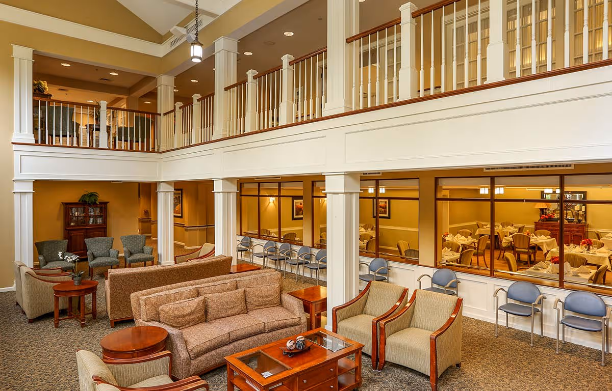 Two-story senior living lounge with sofas, armchairs, coffee tables, and a dining room visible through interior windows.
