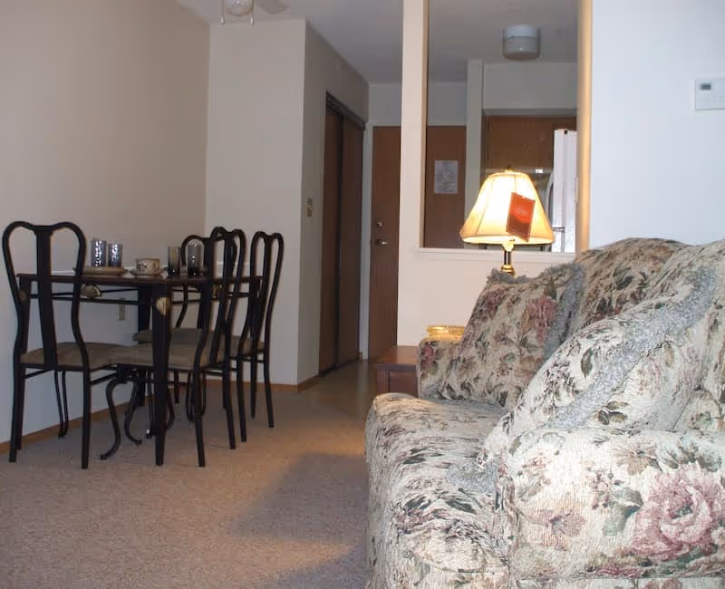 Small apartment living area with a floral couch, a dining table with chairs, and a lamp by the entryway.