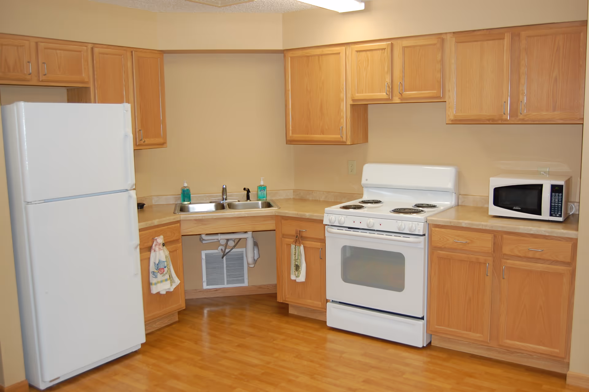 A kitchen area with wooden cabinets, a white refrigerator, a white electric stove with four burners, a microwave on the countertop, a double sink with two soap dispensers, and two hand towels hanging on cabinet handles. The floor is wooden, and the walls are painted beige.