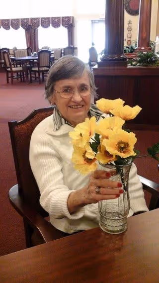 An elderly woman with glasses and short gray hair is sitting at a wooden table in a room with red carpet and large windows. She is smiling and holding a clear glass vase with bright yellow flowers. Behind her are several chairs and tables, and a wooden column with plants on top.