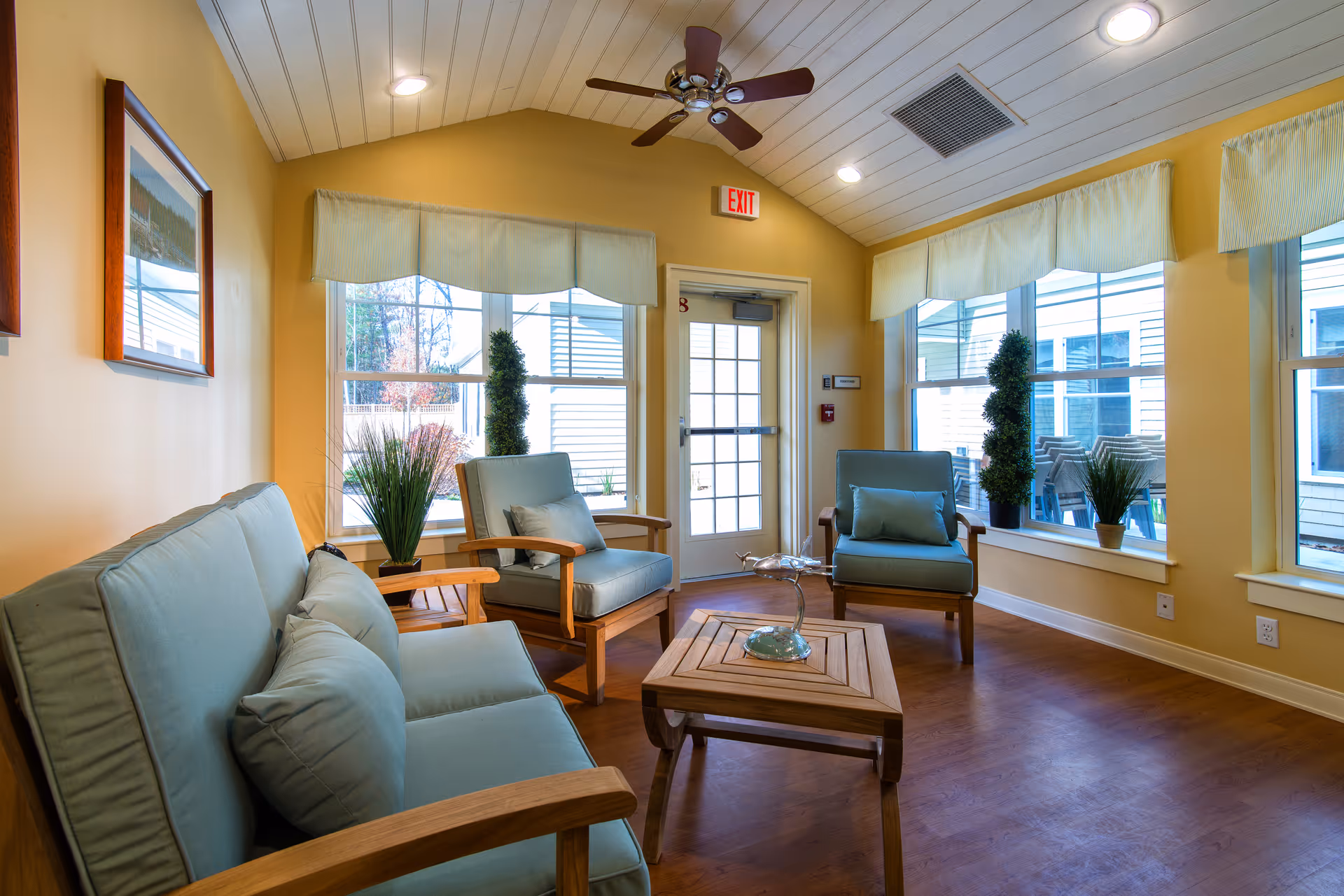 A bright and cozy sitting area with light blue cushioned wooden chairs and a matching loveseat arranged around a wooden coffee table. The room has large windows with light-colored valances, letting in natural light. There are potted plants on the windowsills and a ceiling fan with lights above. The walls are painted a soft yellow, and there is an exit door with a red exit sign above it.