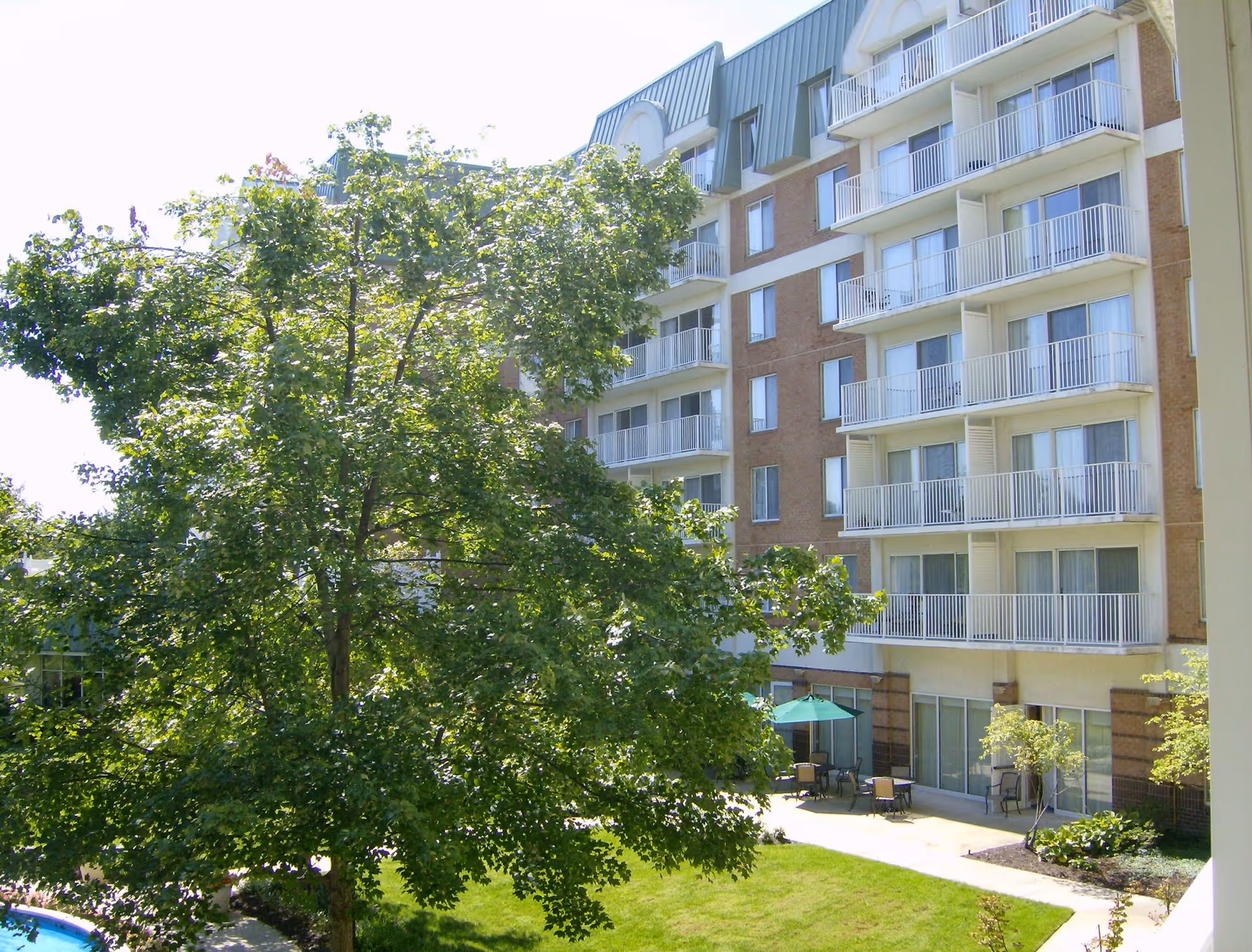 Exterior view of a multi-story residential building with balconies, surrounded by green trees and a small patio area with tables and chairs under an umbrella.