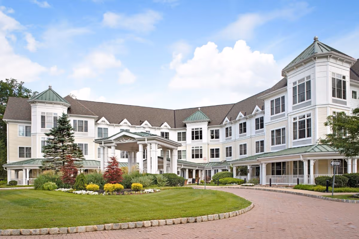 Exterior view of a large, three-story senior living facility building with white siding and green roofs, surrounded by well-maintained landscaping including trees, shrubs, and flowers, under a partly cloudy blue sky.