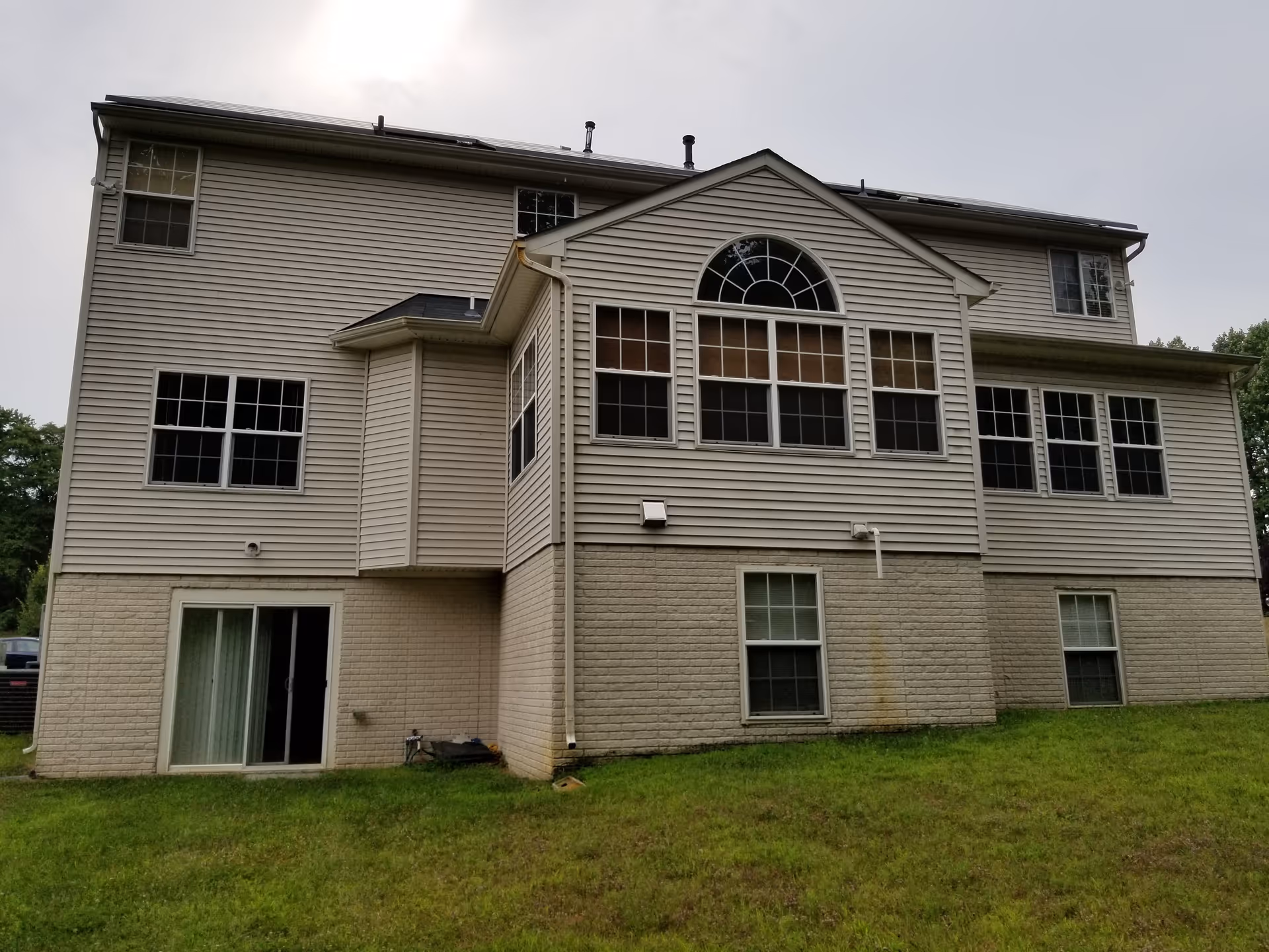Rear exterior view of a two-story house with beige siding and multiple windows, including a large arched window. The house has a sliding glass door on the lower level and a grassy yard in the foreground under an overcast sky.