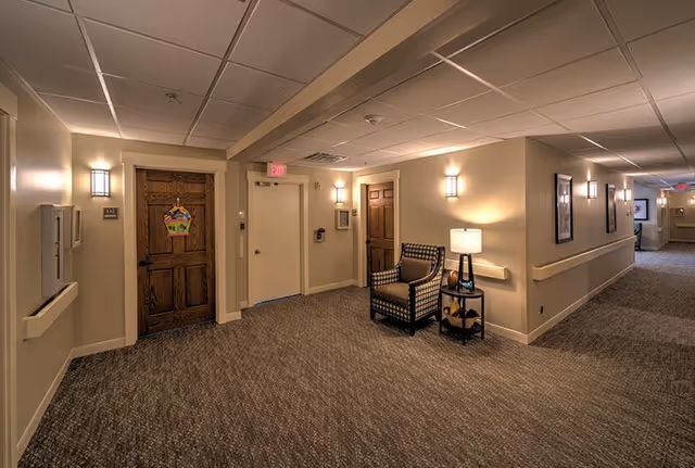 A well-lit hallway in a senior living facility with beige walls, carpeted floor, and wooden doors. There is a single armchair with a patterned cushion next to a small table holding a lamp and decorative items. Wall sconces provide additional lighting, and framed pictures hang on the walls. The hallway extends into the distance with exit signs visible.