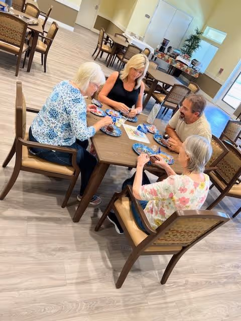 Four adults seated around a table in a communal dining/activity room eating snacks.