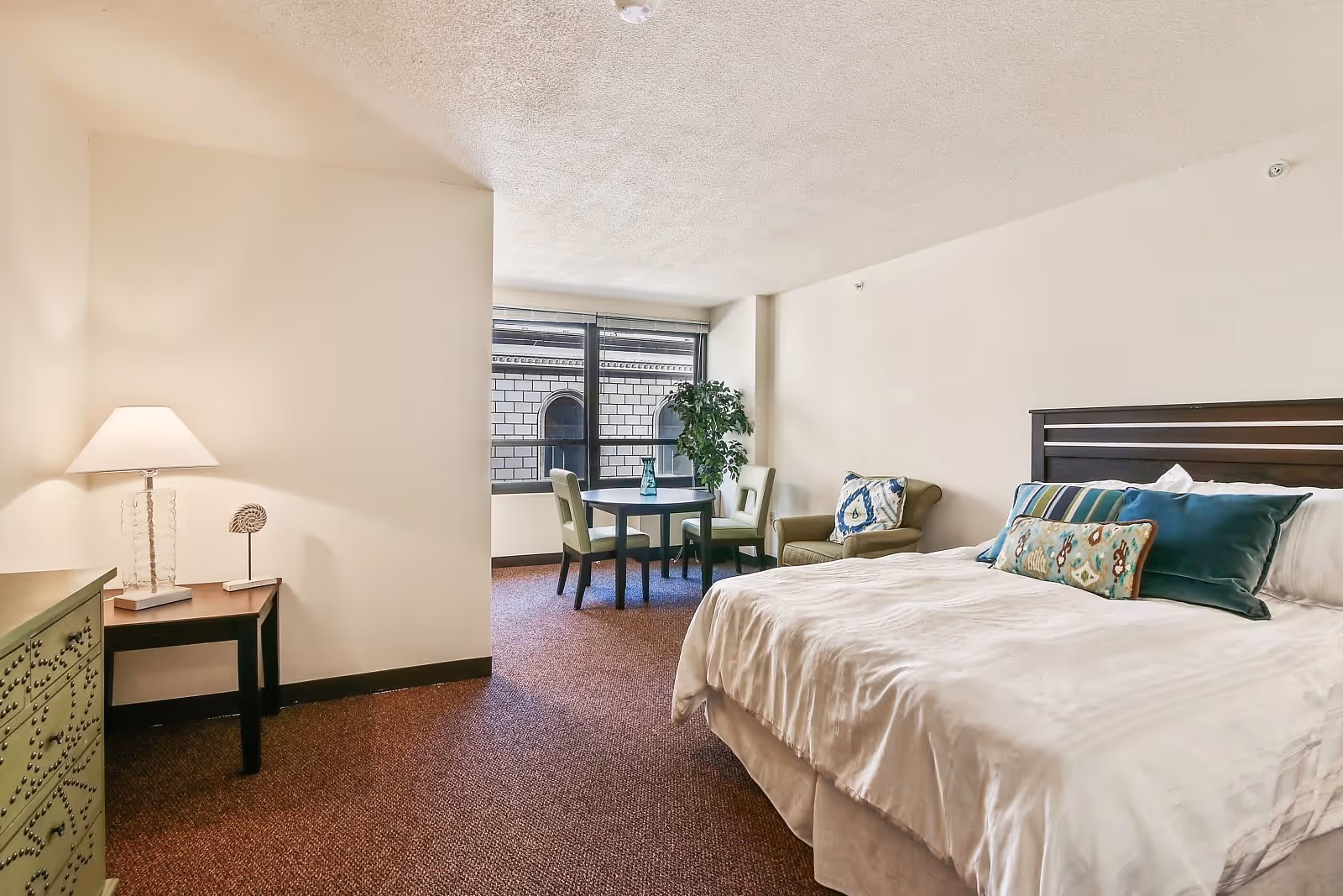 A bright senior apartment bedroom with a large bed adorned with multiple decorative pillows. There is a small round table with two chairs near a window that shows a building exterior. A green armchair with a patterned cushion and a green dresser with a lamp and decorative item are also visible. The room has beige walls and carpeted floor.