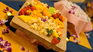 A wooden serving board piled with cubed cheeses and fresh fruit (strawberries, pineapple, mango) garnished with orchid flowers on a decorated table.
