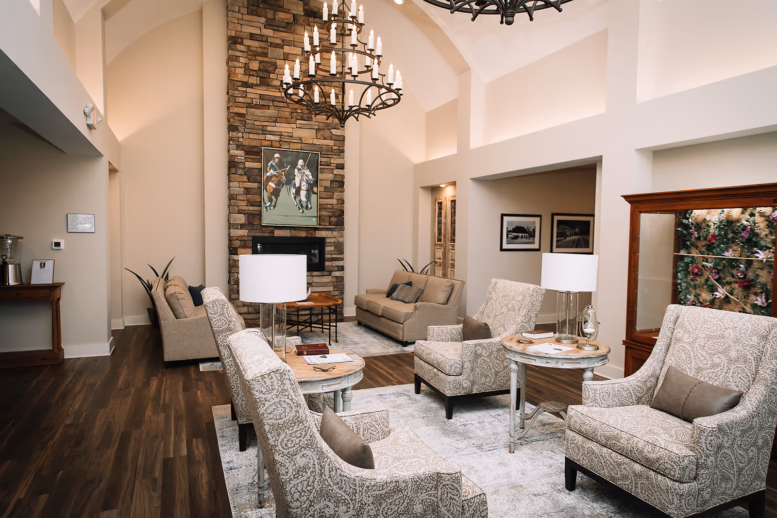 A spacious and elegant living room with high vaulted ceilings, a stone fireplace, and a large painting above it. The room features patterned armchairs, beige sofas, wooden side tables with lamps, and a wooden floor. There is a cabinet with glass doors displaying flowers and framed pictures on the walls.