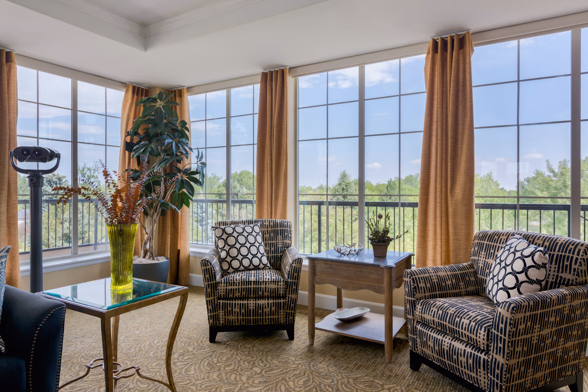 A bright living room area with large windows covered by mustard-colored curtains, two patterned armchairs with circular design pillows, a wooden side table with a small plant and decorative bowl, a glass-top table with a green vase holding dried flowers, and a tall potted plant near the corner. Outside the windows, green trees and a blue sky with some clouds are visible.