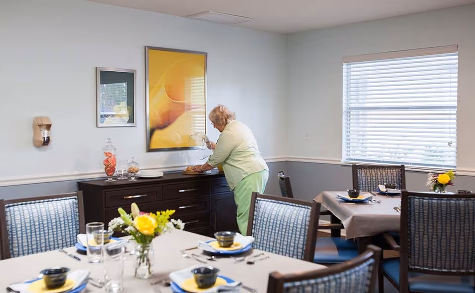 An elderly woman in light green clothing is placing a glass cover over a plate on a dark wooden sideboard in a dining room. The room has several tables set with plates, bowls, glasses, and silverware, each table decorated with a small vase of yellow flowers. The walls are light blue with white trim, and there is a large window with blinds letting in natural light. Two framed pictures hang on the wall above the sideboard.