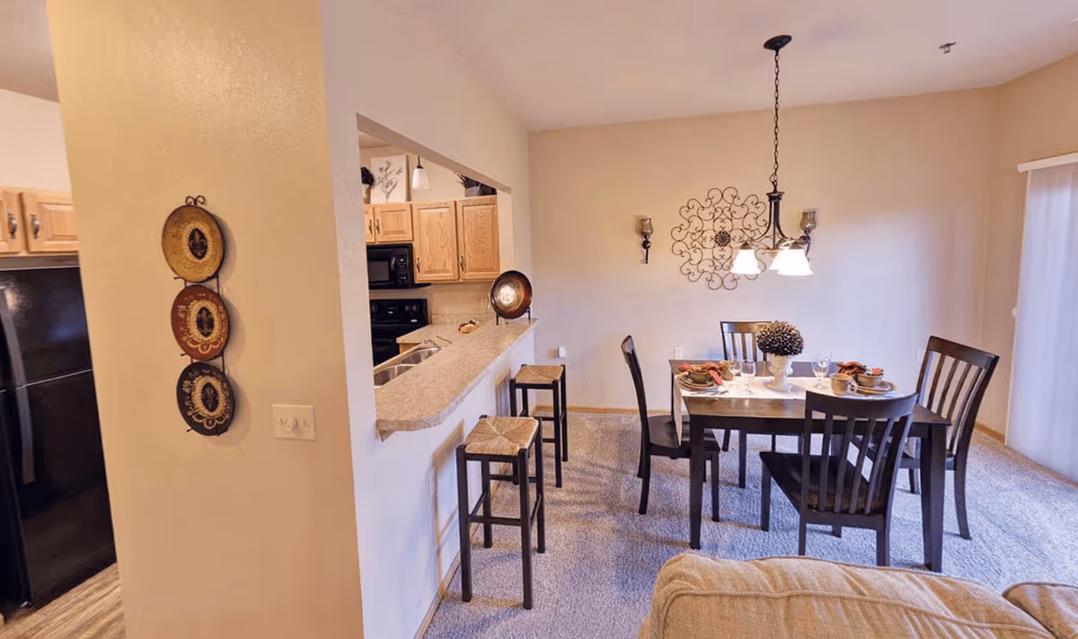 Interior view of a senior living facility dining area with a wooden dining table set for four, dark wooden chairs, a hanging light fixture, and decorative wall art. Adjacent to the dining area is a kitchen with light wood cabinets, a black refrigerator, and a countertop with two bar stools.