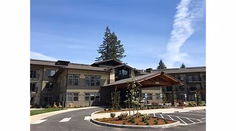 Exterior view of a multi-story senior living facility building with a covered entrance, landscaped garden beds, and a parking area under a blue sky with some clouds.